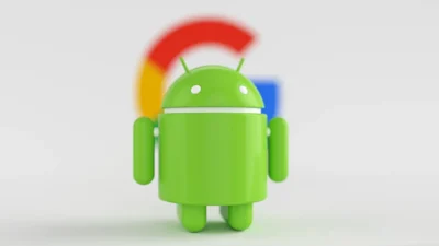 Patung robot Android hijau berdiri di depan logo huruf G Google yang buram di latar putih
