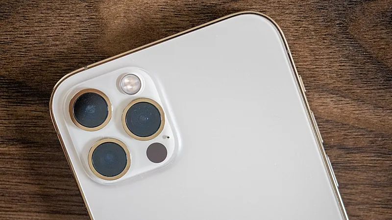 Sudut kiri atas punggung iPhone 12 Pro warna perak: tiga lensa kamera melingkar berbingkai emas, lampu kilat, dan satu titik gelap sensor LiDAR, di atas meja kayu