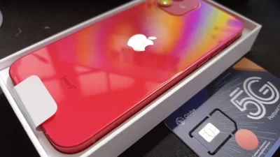 Punggung iPhone 12 warna merah PRODUCT RED masih berbaring di dalam kotak putihnya, bersebelahan dengan kartu SIM 5G
