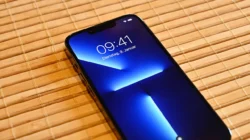 iPhone 13 Pro menghadap atas di atas tikar bambu, layar menyala menampilkan jam 09.41 dan wallpaper biru