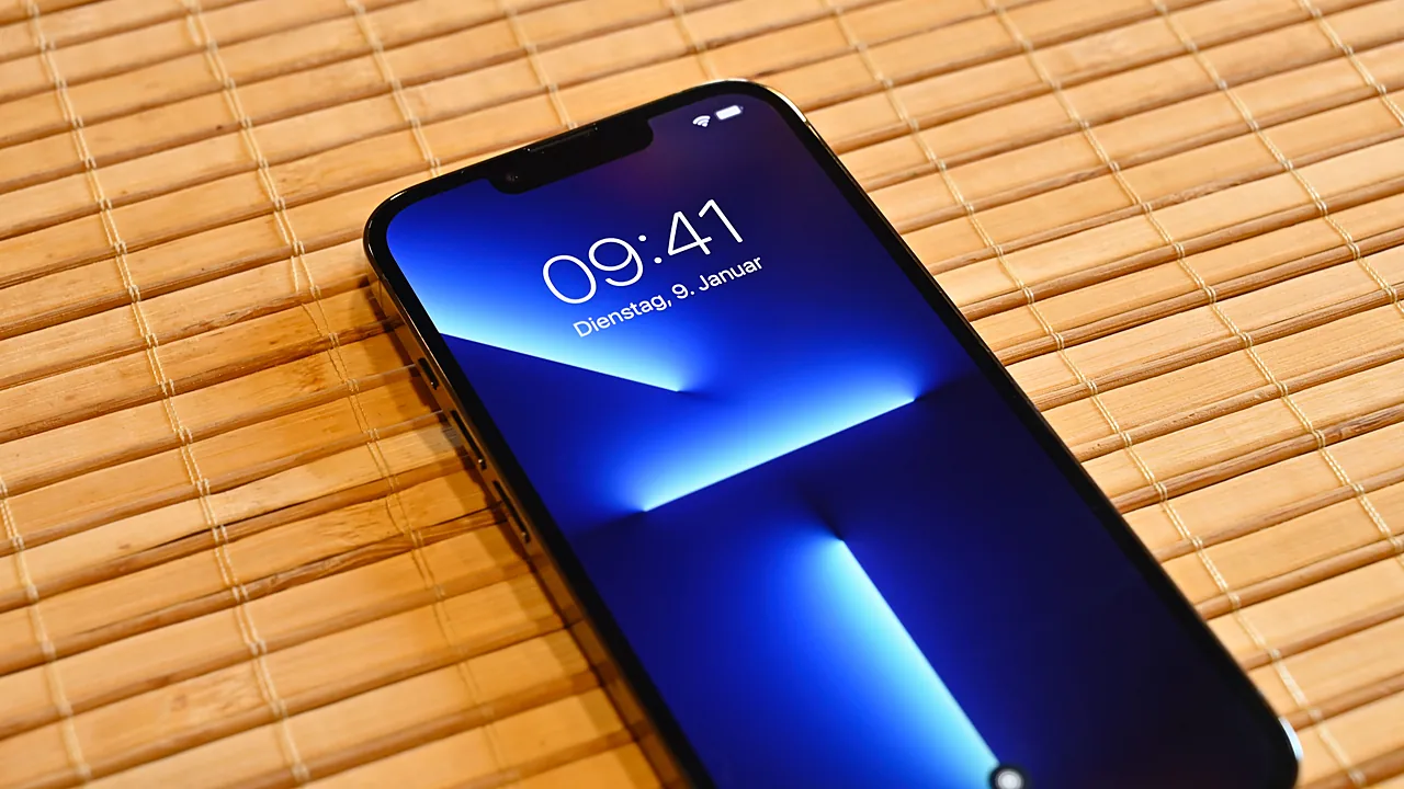 iPhone 13 Pro menghadap atas di atas tikar bambu, layar menyala menampilkan jam 09.41 dan wallpaper biru