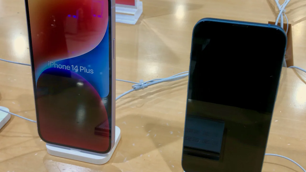 Unit pajang iPhone 14 Plus menyala menampilkan namanya di layar, berdiri di samping iPhone 14 warna biru yang layarnya mati, di meja kayu gerai Apple