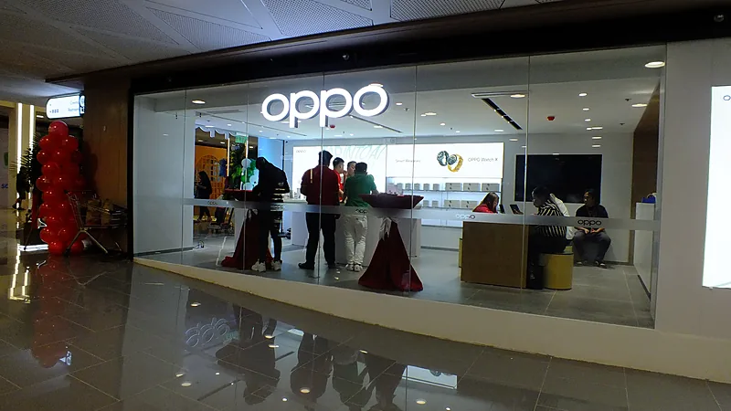 Gerai resmi OPPO di dalam mal dengan papan nama OPPO menyala putih di atas kaca depan, beberapa pengunjung berdiri di meja pajangan, dan dua petugas melayani di meja kasir sebelah kanan