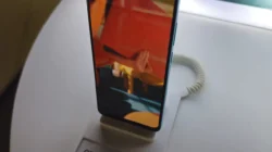 OPPO A78 berdiri di dudukan pajangan toko, layarnya menyala menampilkan gambar berwarna oranye dan biru, dengan kabel pengaman spiral putih terpasang di belakangnya