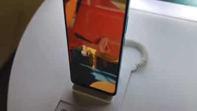 OPPO A78 berdiri di dudukan pajangan toko, layarnya menyala menampilkan gambar berwarna oranye dan biru, dengan kabel pengaman spiral putih terpasang di belakangnya