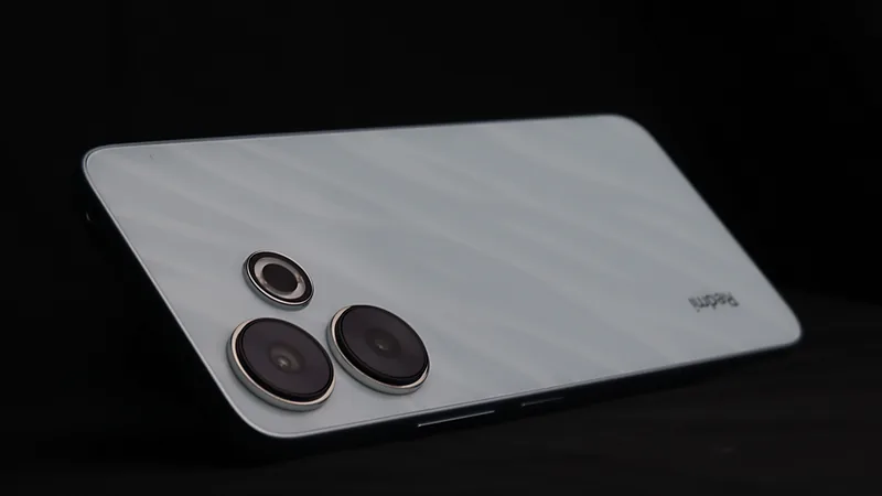 Punggung Redmi 13 berwarna putih keperakan tergeletak miring di atas permukaan hitam, memperlihatkan tiga lensa kamera bulat di pojok kiri atas dan tulisan Redmi di sisi kanan bawah