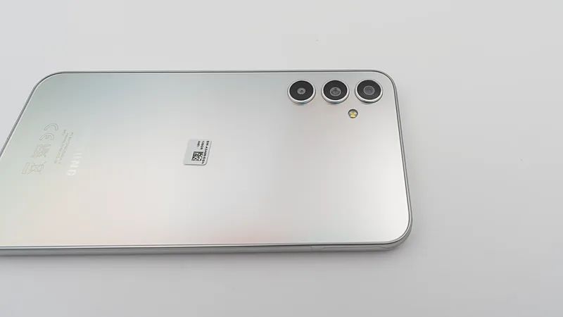 Punggung Samsung Galaxy A34 5G warna perak tergeletak miring di atas permukaan putih, memperlihatkan tiga lingkaran kamera dan lampu kilat kuning di sudut kanan atas serta label regulasi kecil di tengah bodi