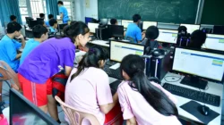 Belasan siswa SMP duduk berderet menghadap komputer di sebuah ruang kelas komputer, sebagian memakai headset
