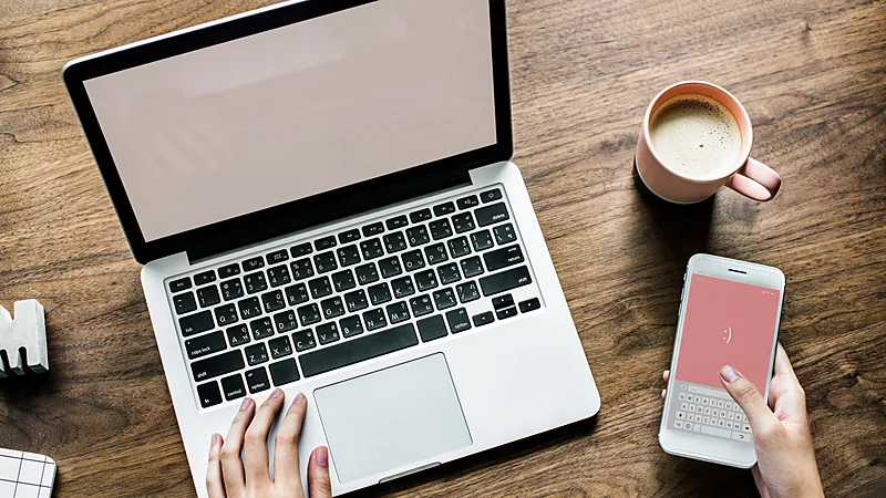 Laptop terbuka, ponsel dengan papan ketik di layar, dan secangkir kopi di meja kayu dilihat dari atas