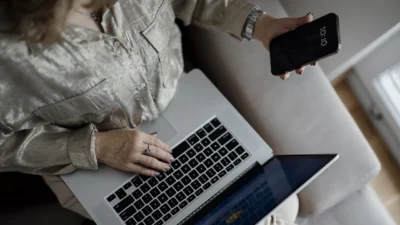 Perempuan duduk di sofa mengetik di laptop sambil memegang ponsel di tangan kanan