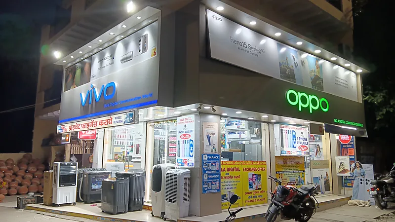 Gerai HP bertanda vivo dan OPPO menyala pada malam hari dengan papan berbahasa Hindi di Dabri, Delhi